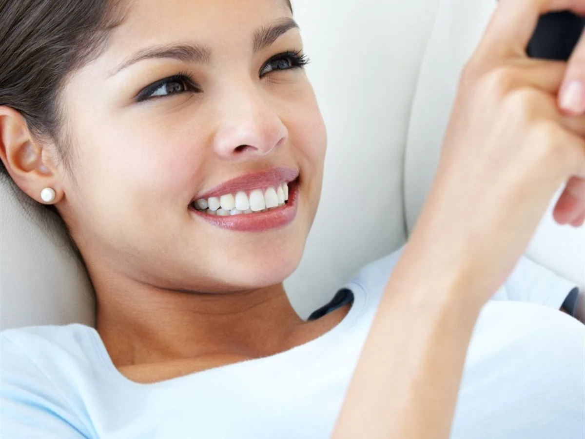 A woman relaxing back on a sofa, smiling at her phone as she messages a friend.